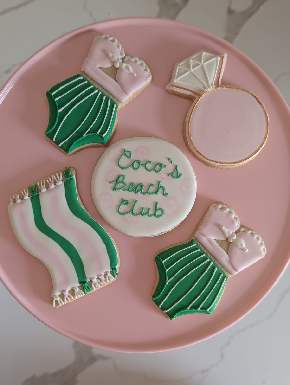 Beach club bachelorette cookies on a pink cake stand - green and white striped swimsuit cookies with pink tops, a pink and green striped beach towel with fringe, a diamond ring cookie, and a round cookie hand-lettered in green script