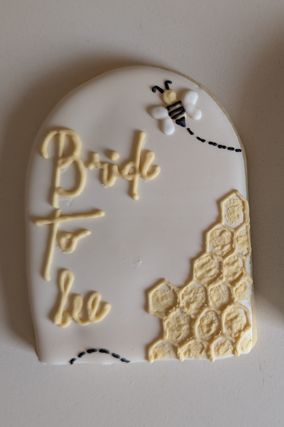 An arch-shaped bridal shower cookie in ivory fondant lettered Bride to be in butter yellow script, with a piped honeycomb corner and a little bee following a dashed flight path