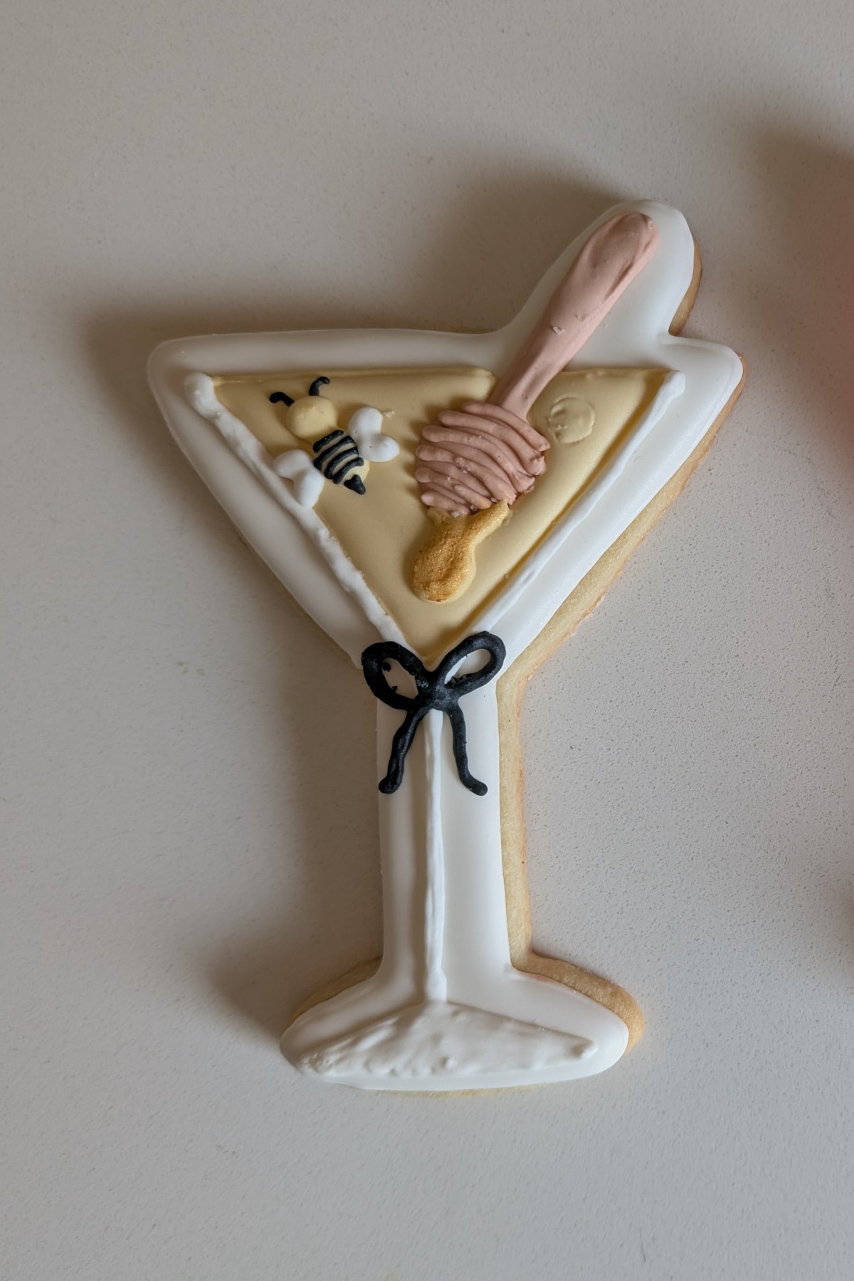 A martini glass cookie iced in white and honey yellow with a honey dipper, a bee and a small black bow tied around the stem