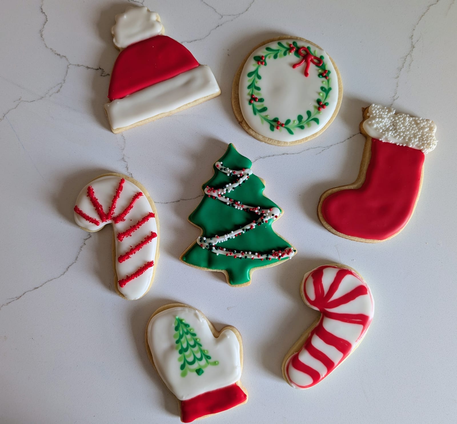 Seven decorated Christmas sugar cookies on white marble - a green tree with sprinkle garlands, a red stocking, a santa hat, a white mitten, a holly wreath and two candy canes