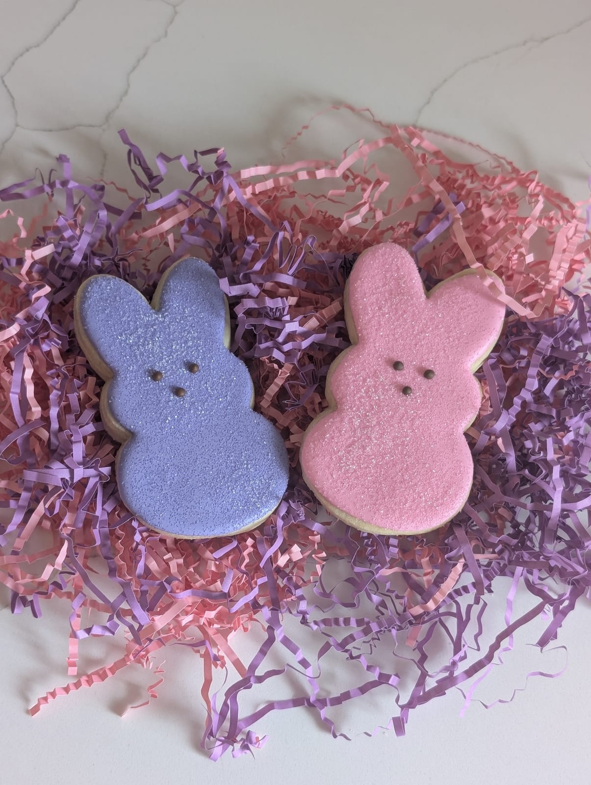 A pink and a lavender bunny-shaped sugar cookie coated in sparkling sanding sugar, sitting on a nest of pink and purple shredded paper Easter grass
