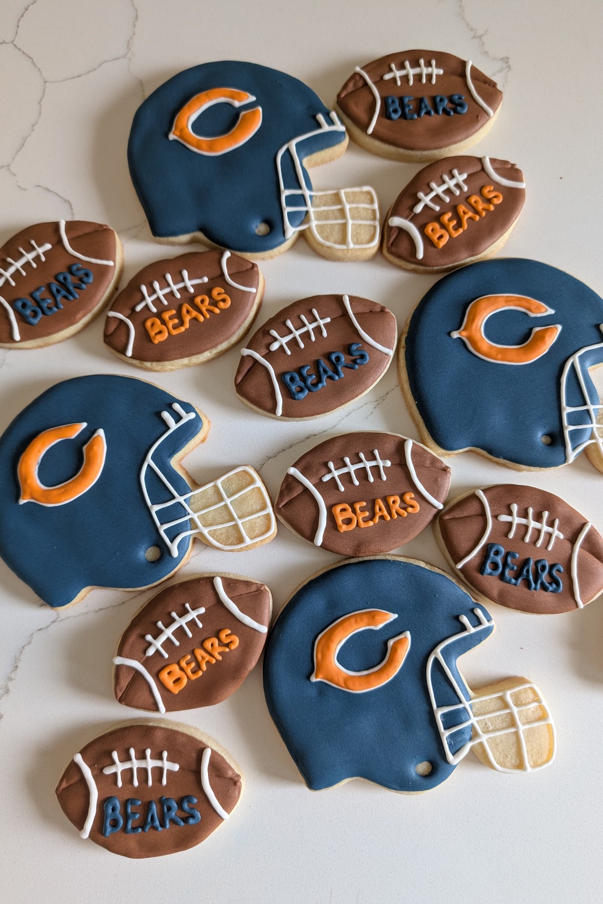 A game day cookie set on white marble - navy football helmet cookies with an orange letter, and brown football cookies with white laces lettered in orange and navy