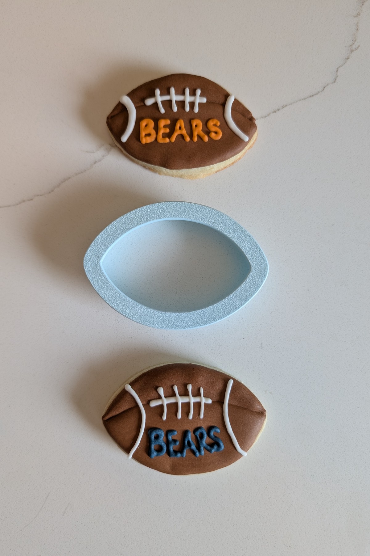 Two decorated football cookies in brown royal icing with piped white laces, either side of the pale blue 3D printed football cookie cutter that cut them