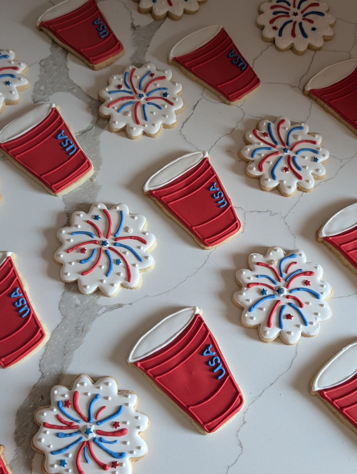 Fourth of July cookies - scalloped white rounds piped with red and blue firework bursts and star sprinkles, alternating with red party cup cookies lettered USA in blue
