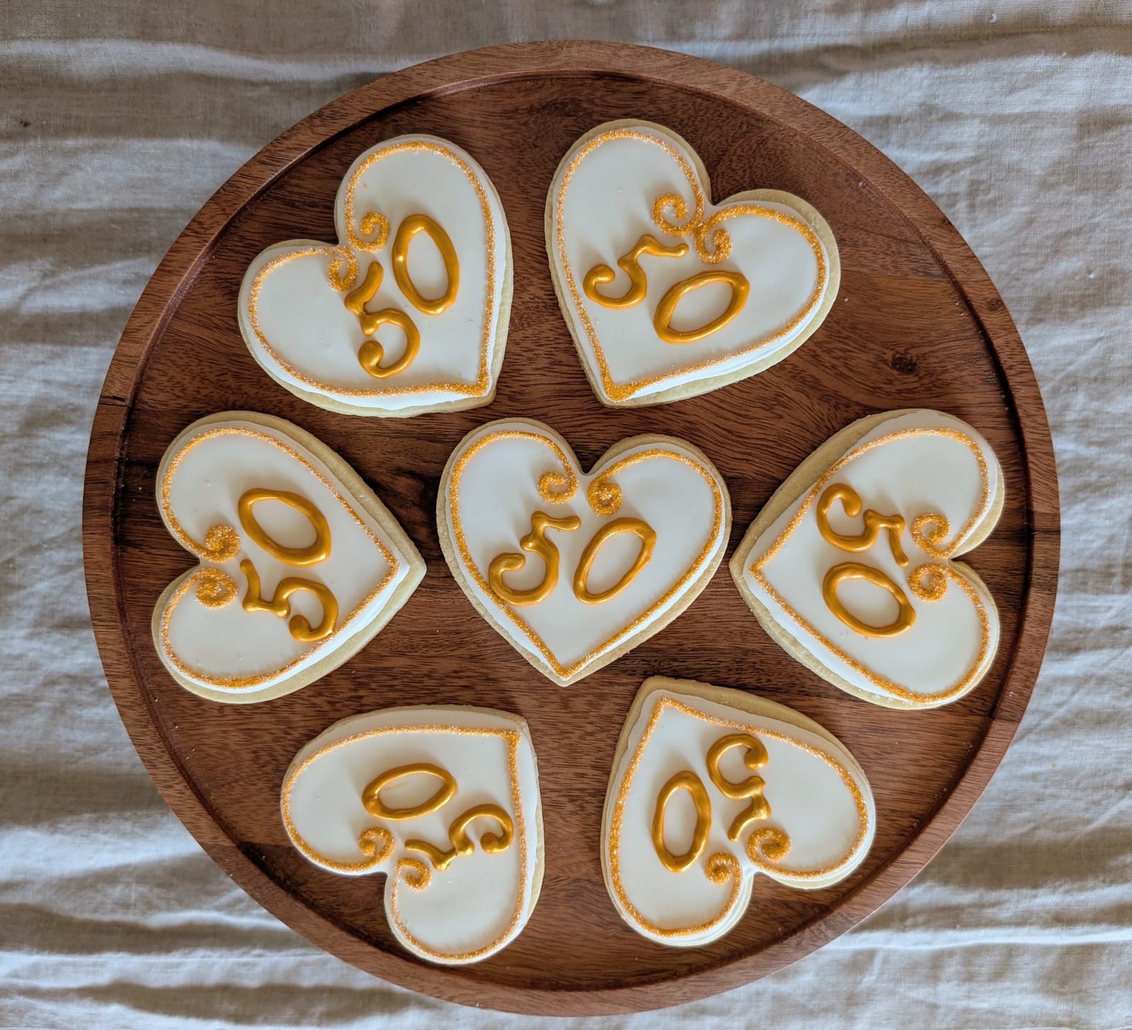 Seven ivory heart-shaped cookies arranged on a round walnut board, each edged in gold sanding sugar and piped with a gold number fifty in a flourished script
