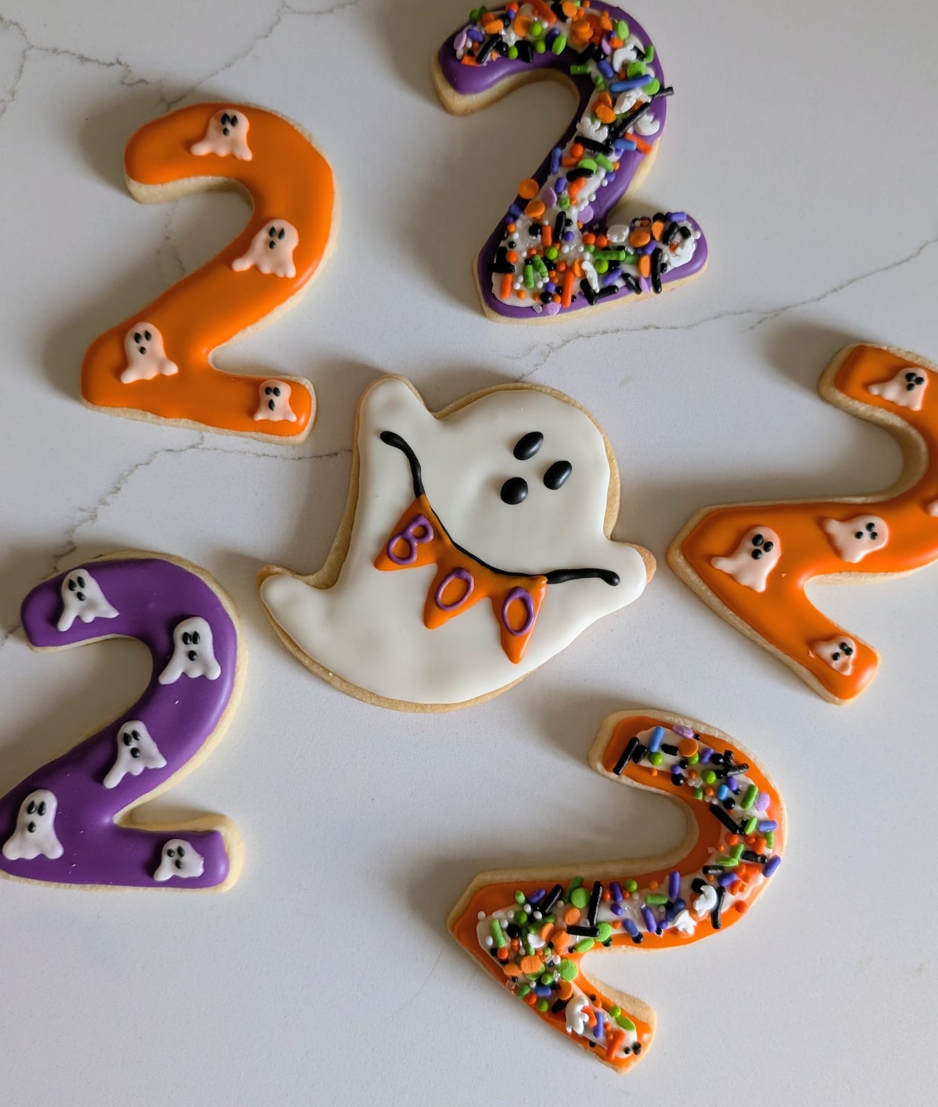 Halloween birthday cookies - a white ghost holding an orange BOO banner, surrounded by orange and purple number two cookies decorated with tiny ghosts and Halloween sprinkles