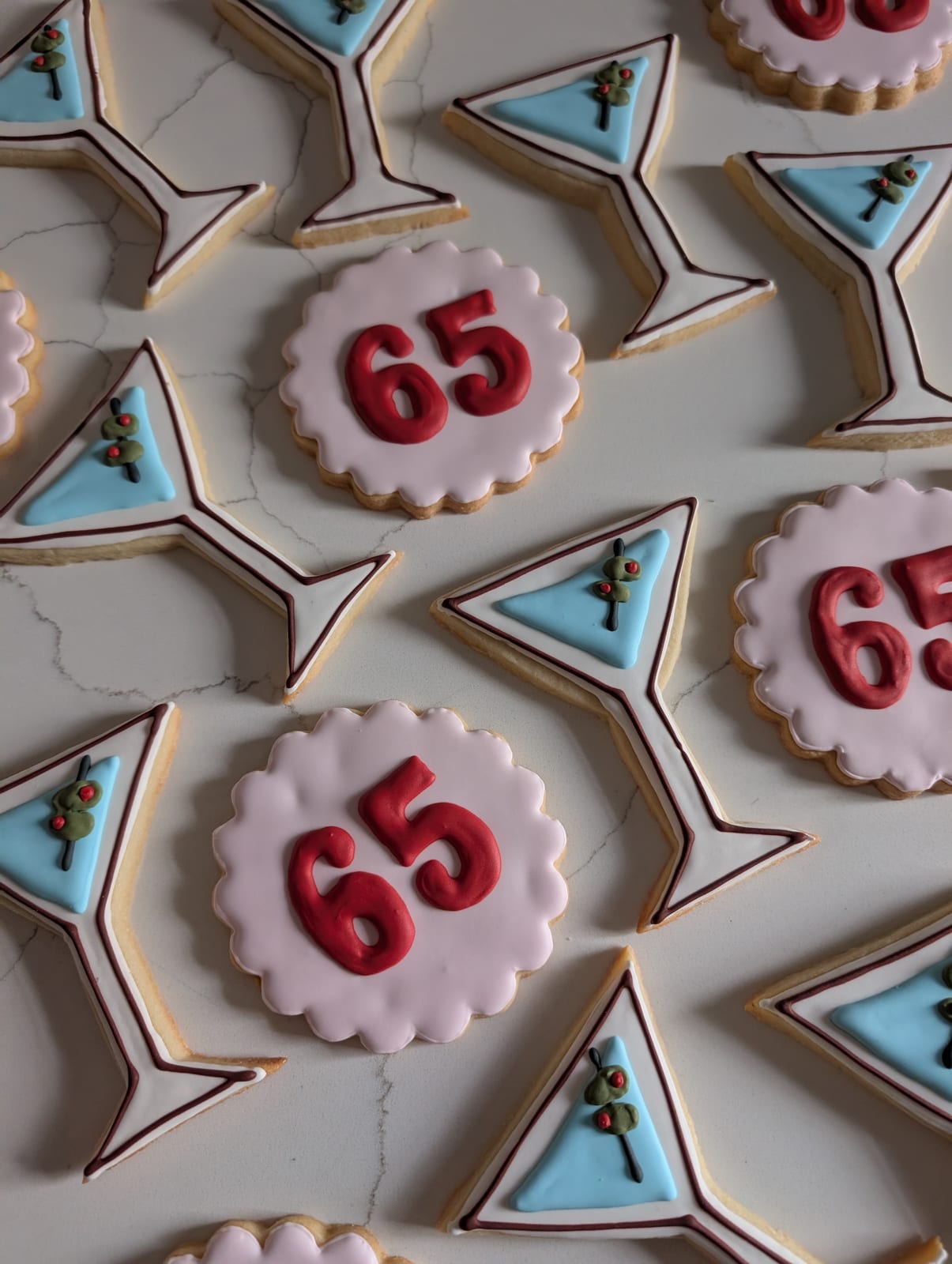Cocktail themed birthday cookies - martini glass cookies iced white with pale blue drinks and green olives on a pick, alternating with scalloped pink rounds piped with a red 65