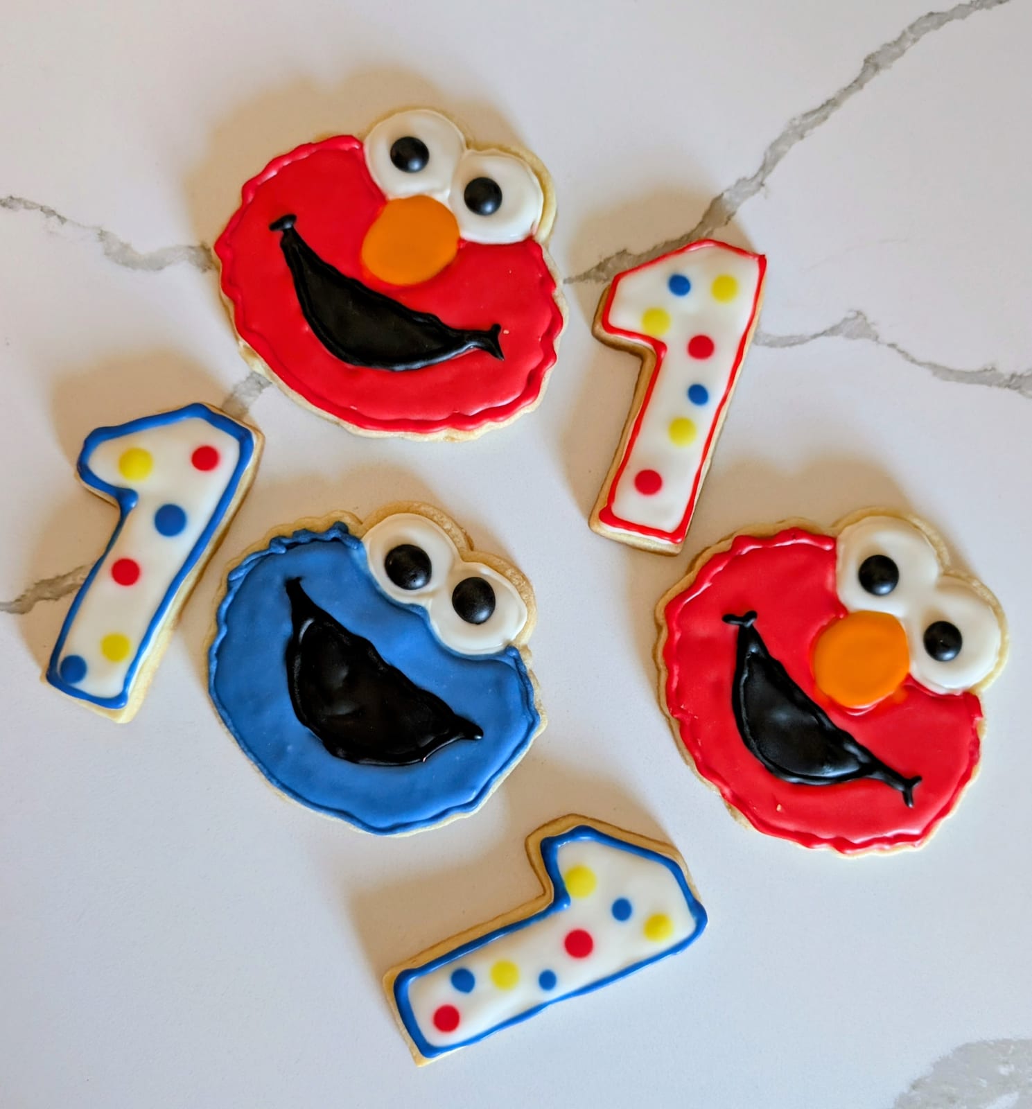 First birthday cookies on white marble - two round red monster faces with big white googly eyes, an orange nose and an open smiling mouth, a blue monster face with the same eyes, and white number one cookies dotted in red, yellow and blue