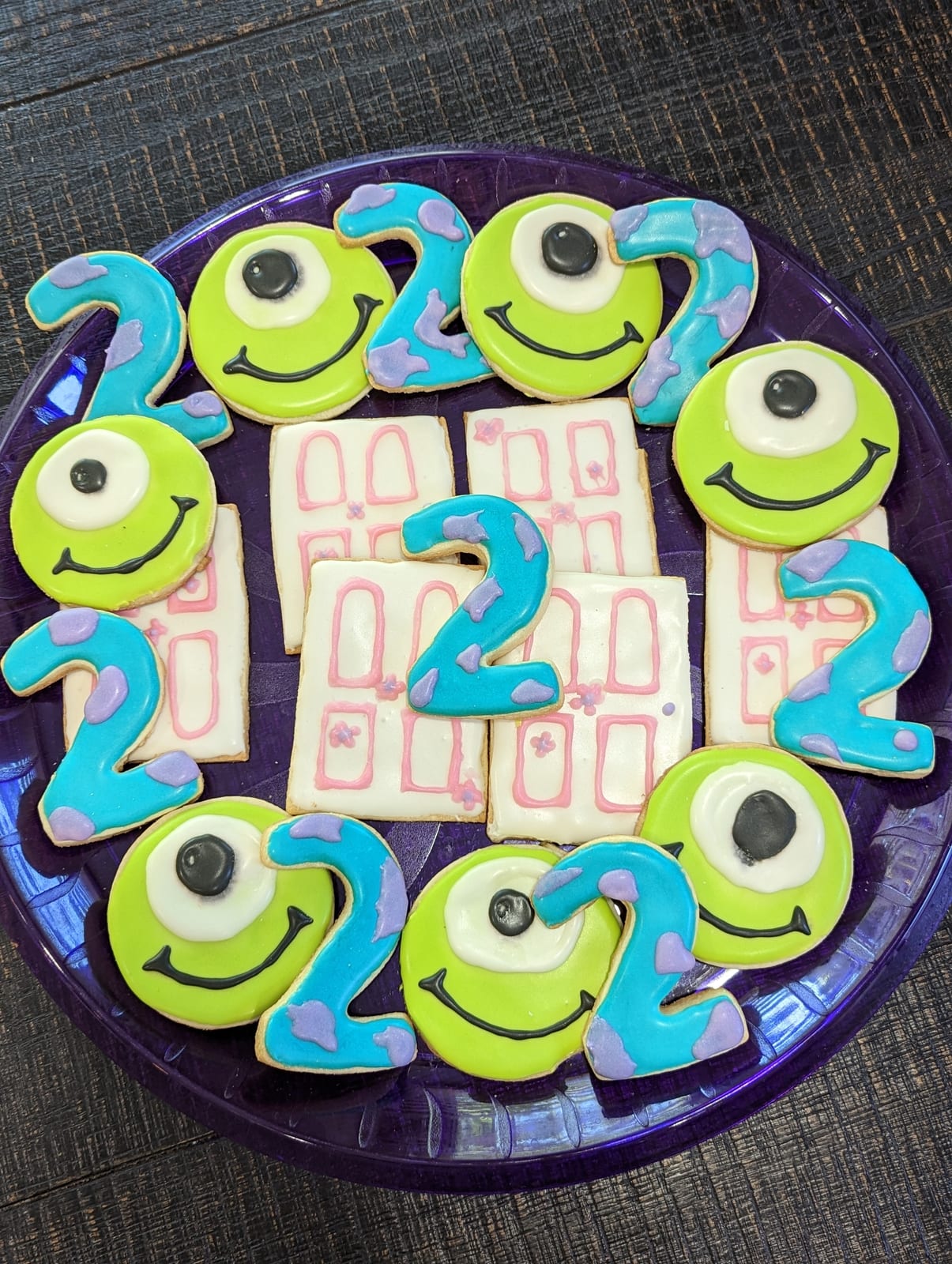 Monster themed second birthday cookies arranged on a purple plate - round lime green monster faces with one large eye and a wide smile, blue and lavender spotted number two cookies, and white rectangular door cookies outlined in pink