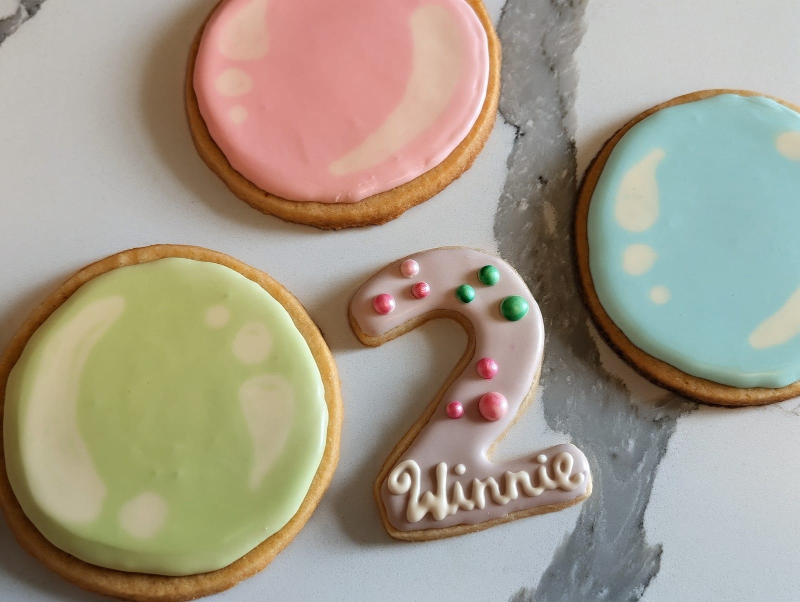 Pastel second birthday cookies - glossy round cookies iced in pink, soft green and pale blue, beside a pale pink number two cookie set with pink and green metallic sugar pearls and piped with a name
