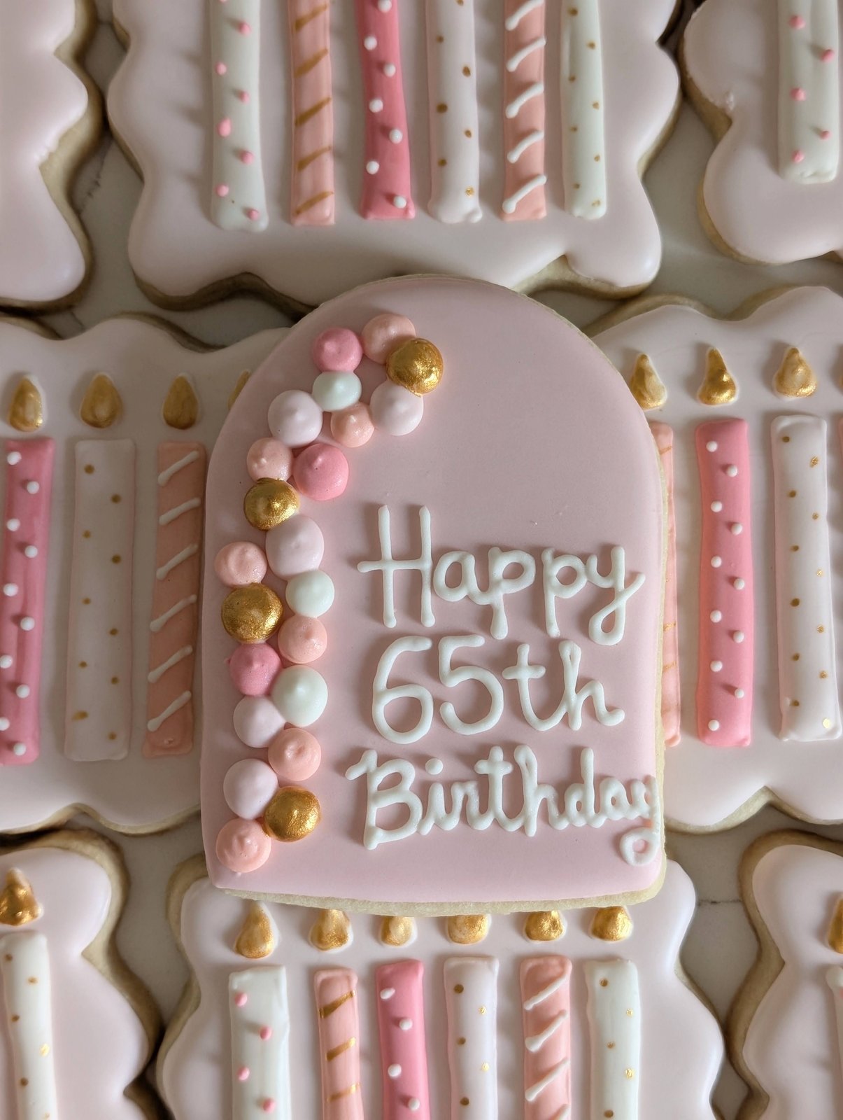 A pink Happy 65th Birthday plaque cookie with a piped balloon garland in pink, white and gold, resting on a bed of birthday candle cookies in blush, pink and gold