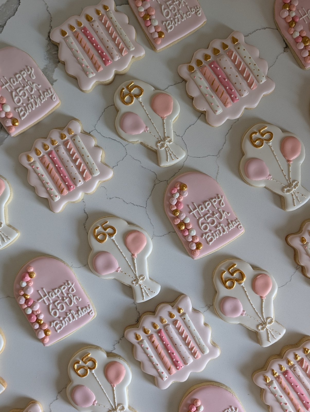 A pink and gold 65th birthday cookie set on white marble - Happy 65th Birthday arch plaques, scalloped cookies piped with rows of birthday candles, and white balloon bouquet cookies with a gold 65