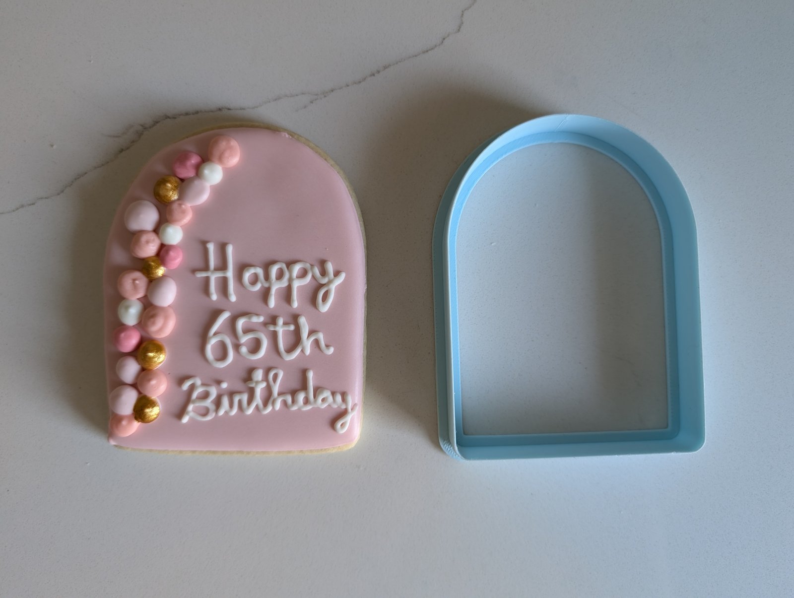 A Happy 65th Birthday arch plaque cookie in pink royal icing beside the blue 3D printed arch cookie cutter that cut it