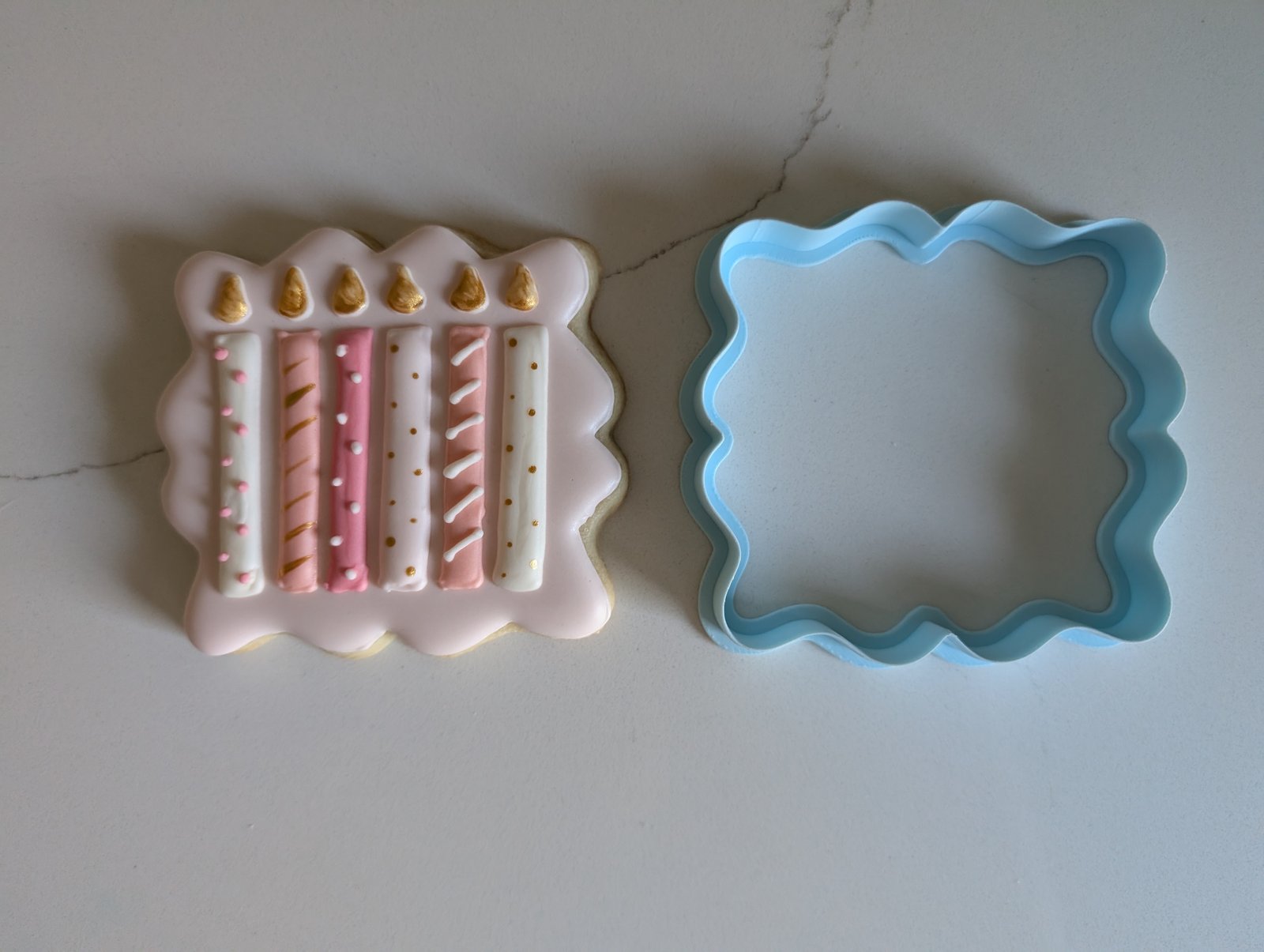 A scalloped plaque cookie piped with six pink, white and rose birthday candles with gold flames, beside its blue 3D printed wavy plaque cookie cutter