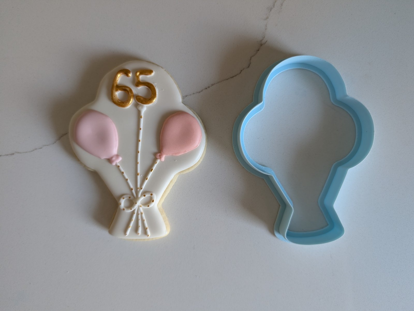 A white balloon bouquet cookie with two pink balloons tied in a bow and a gold 65, beside the blue 3D printed balloon bouquet cookie cutter that cut it