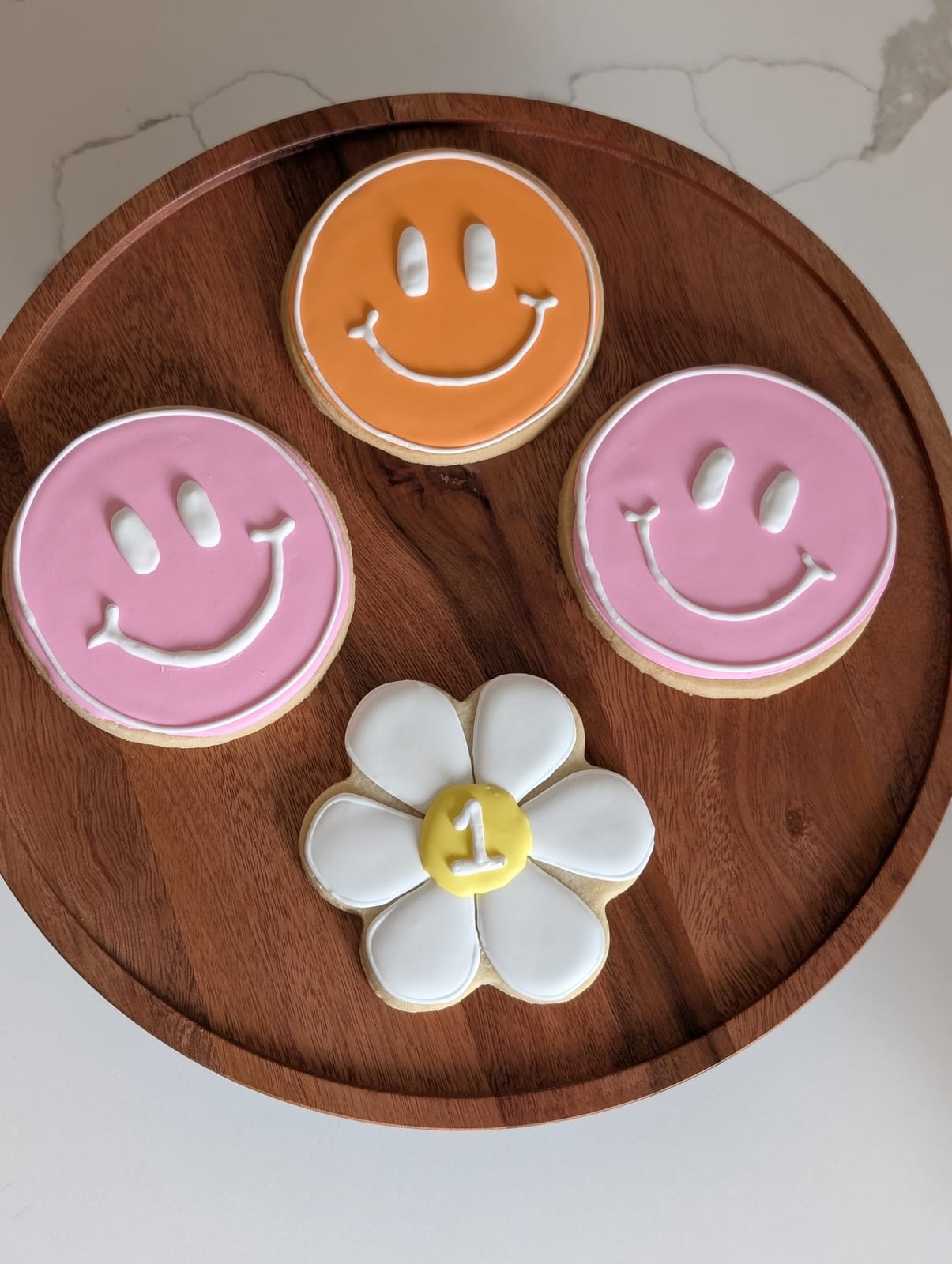 Retro smiley face cookies in pink and orange with white piped features, beside a white daisy cookie with a yellow centre piped with the number one, on a round walnut board