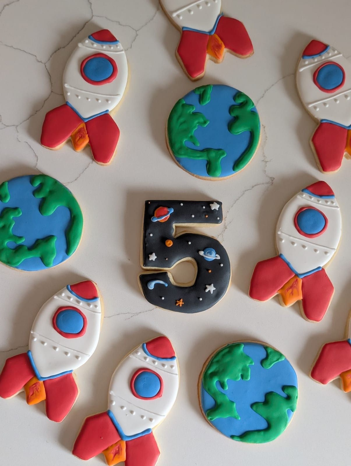 Space themed birthday cookies - white rocket cookies with red fins and orange flames, round earth cookies iced blue and green, and a black number five cookie painted with stars and planets