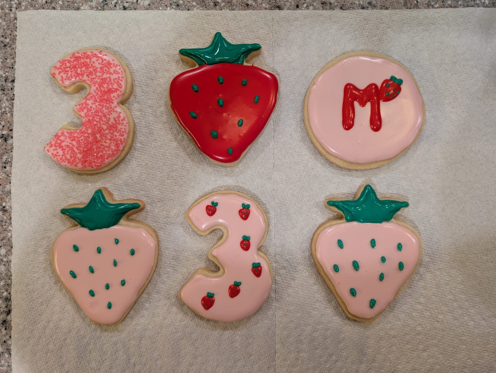 Six strawberry-themed birthday cookies - a red strawberry and two blush pink ones with green piped hulls and seeds, two number three cookies in pink, and a round pink cookie with a red piped monogram