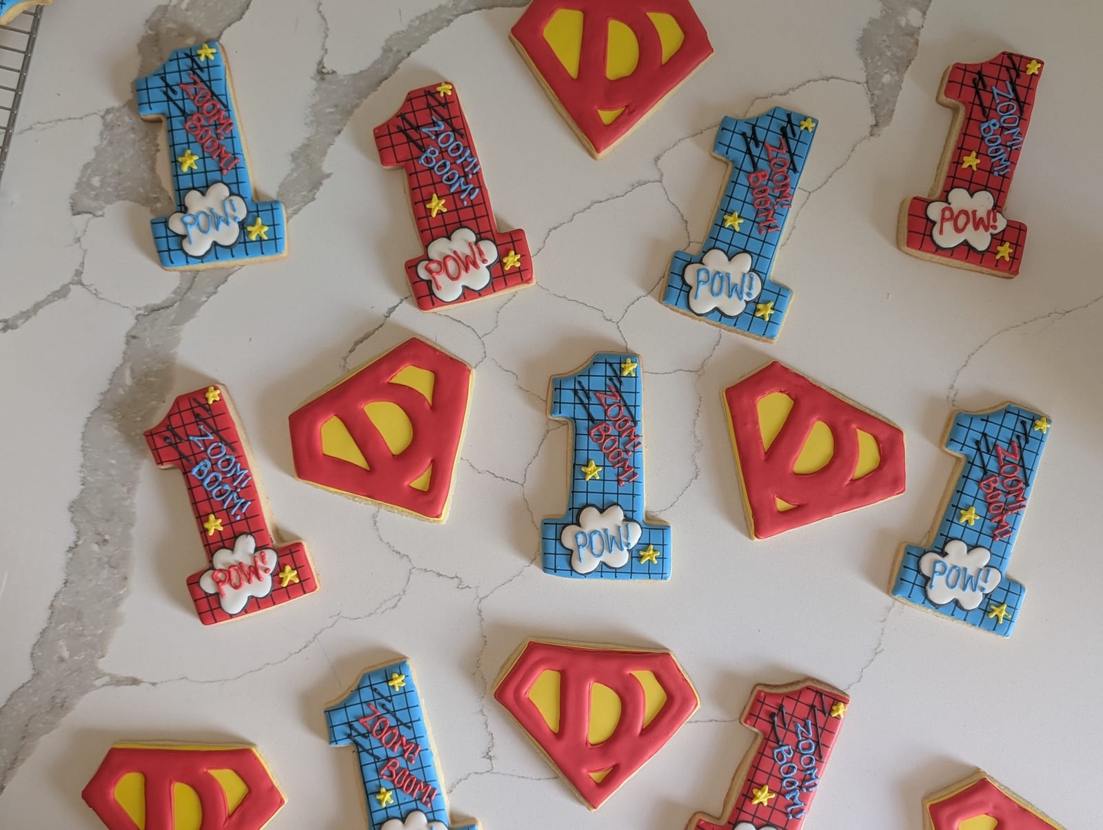 Superhero first birthday cookies on white marble - red and blue number one cookies iced in a comic-book halftone grid and lettered ZOOM, BOOM and POW in speech bubbles, alternating with red and yellow shield cookies monogrammed with a D