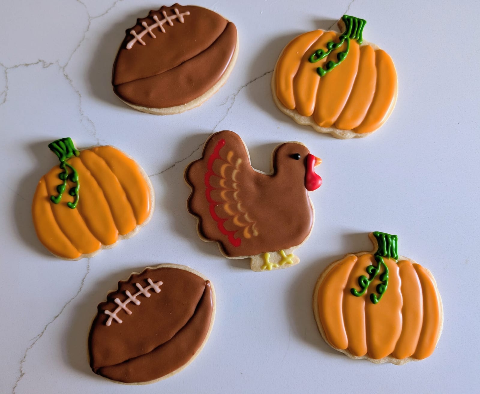 Six Thanksgiving cookies on white marble - a brown turkey with red and cream tail feathers, three ribbed orange pumpkins with green stems, and two brown footballs with white laces