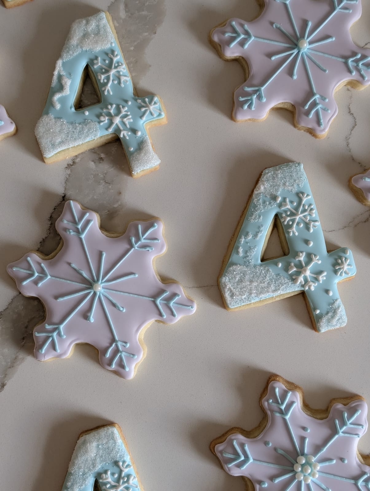 Winter birthday cookies - pale pink snowflake cookies piped with ice blue crystal patterns, beside pale blue number four cookies topped with white sanding sugar snow